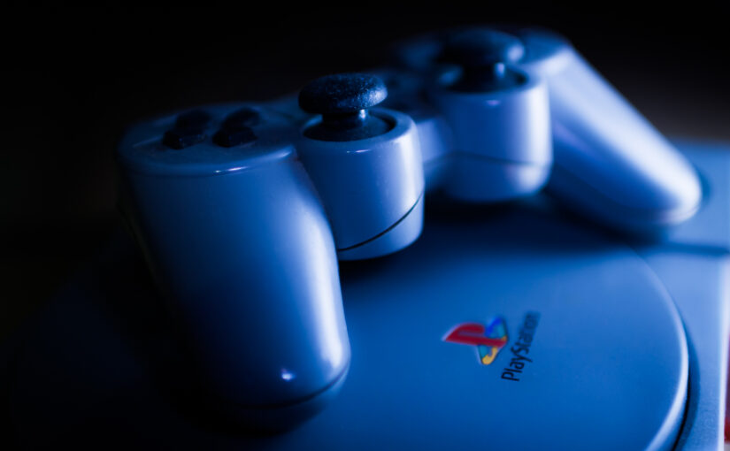 A moodily lit photograph of an original playstation with the playstation logo visible on the disc drive, with its controller place on top.